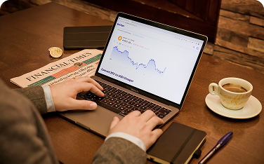 Overhead view of a person's hands typing on a laptop that displays a financial market chart. A newspaper labeled "FINANCIAL TIMES" and a cup of coffee are also on the wooden table.