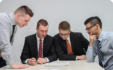 Four young men in business attire are gathered around a small table, intently focused on reviewing a document placed between them.