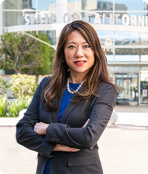 Fiona Ma, California State Treasurer, stands with her arms crossed, smiling in a gray pinstripe suit and pearls, with a modern State of California building visible behind her.